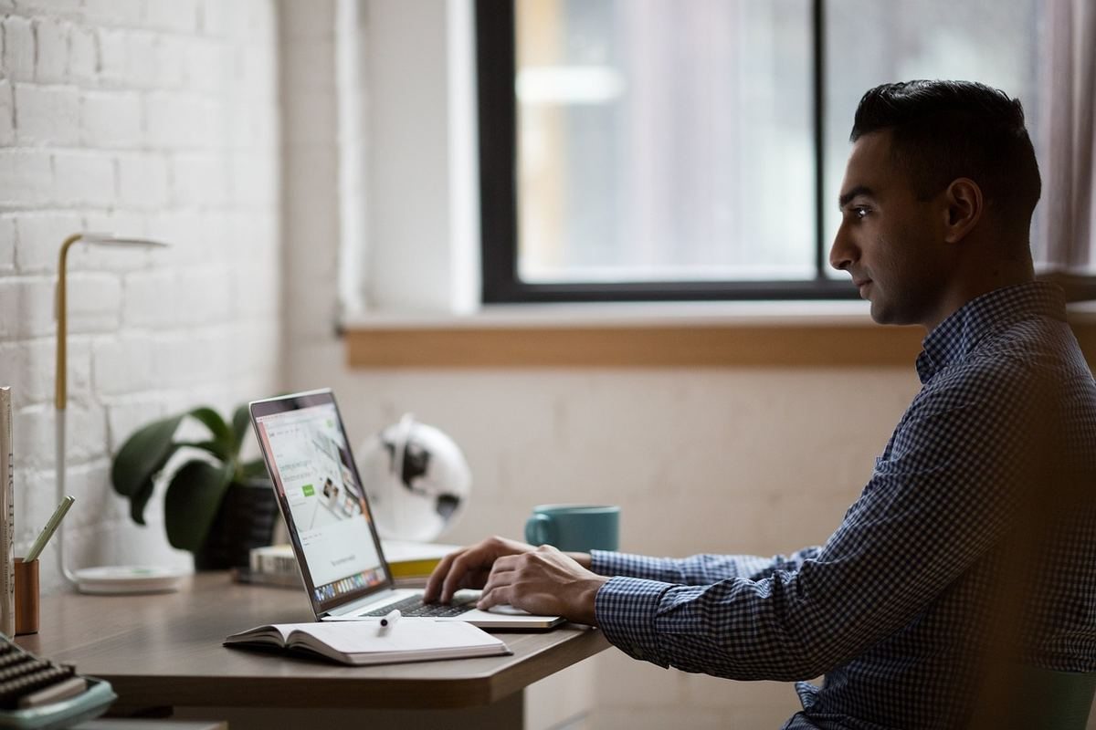 Homme en chemise bleue travaillant sur ordinateur portable à un bureau avec plante et tasse de café