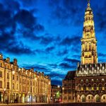 Vue nocturne d'une place avec un clocher illuminé et façades anciennes