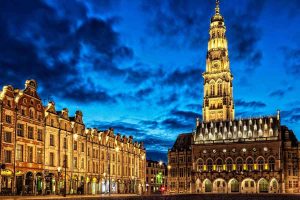 Vue nocturne d'une place avec un clocher illuminé et façades anciennes